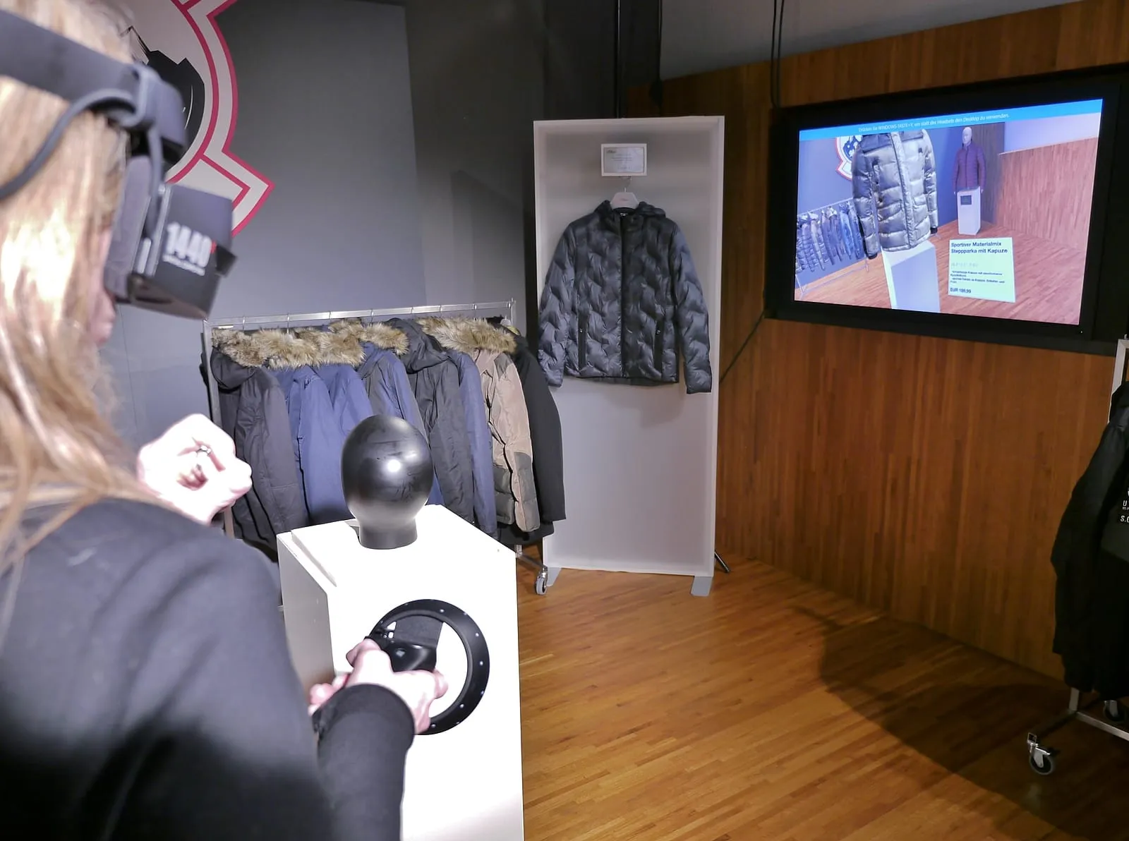 Nutzerin mit VR-Headset erkundet den virtuellen s.Oliver Showroom – physische Jacken im Hintergrund, Bildschirm zeigt die VR-Ansicht mit Produktdetails