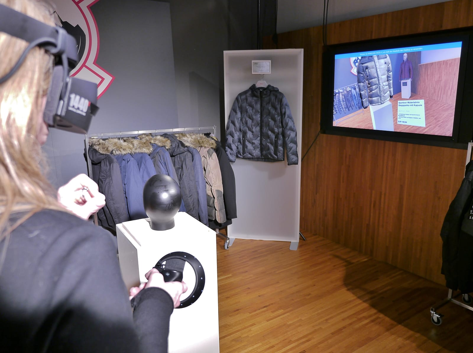Nutzerin mit VR-Headset erkundet den virtuellen s.Oliver Showroom – physische Jacken im Hintergrund, Bildschirm zeigt die VR-Ansicht mit Produktdetails