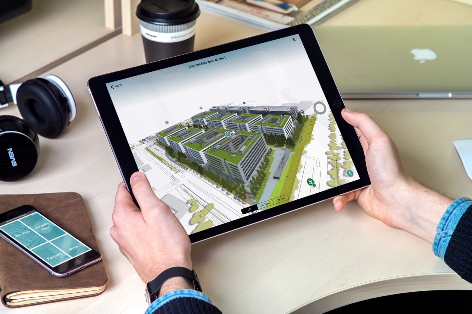 iPad zeigt interaktives 3D-Modell des Siemens Campus Erlangen Modul 1 mit Gebäuden und Grünflächen – iPhone mit App-Menü im Hintergrund